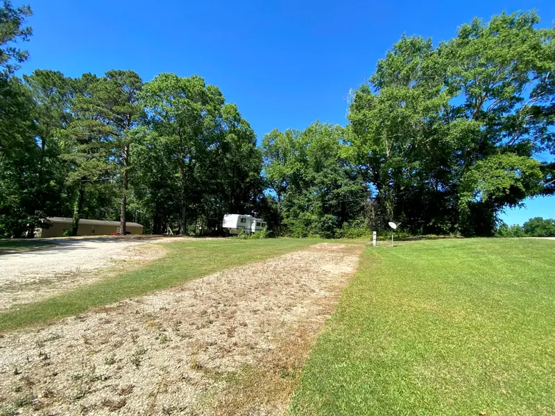 A gravel lot beside a mown grass lane, with a camper under the trees beyond