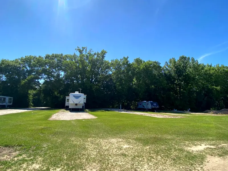 A fifth-wheel camper on a gravel pad backed up to the trees, with open grass around it
