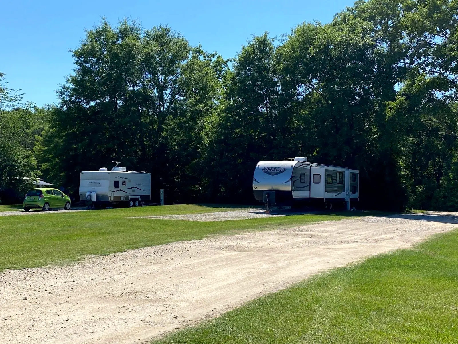 Two travel trailers on gravel lots at the tree line, with an open grass field in front